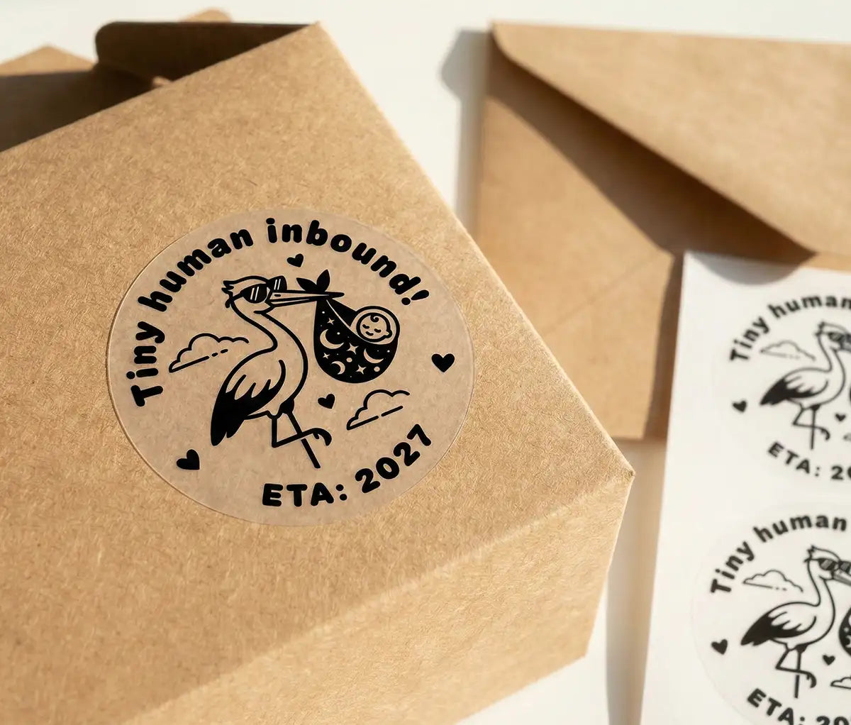 A stork carrying a baby, and the text "Tiny human inbound!" with a custom year is printed in black on a transparent sticker. The sticker is applied to a Kraft box.