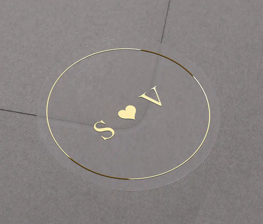 Gold foil wedding sticker with custom initials "S and V" and a small heart centered within it. The label is applied to a grey envelope.