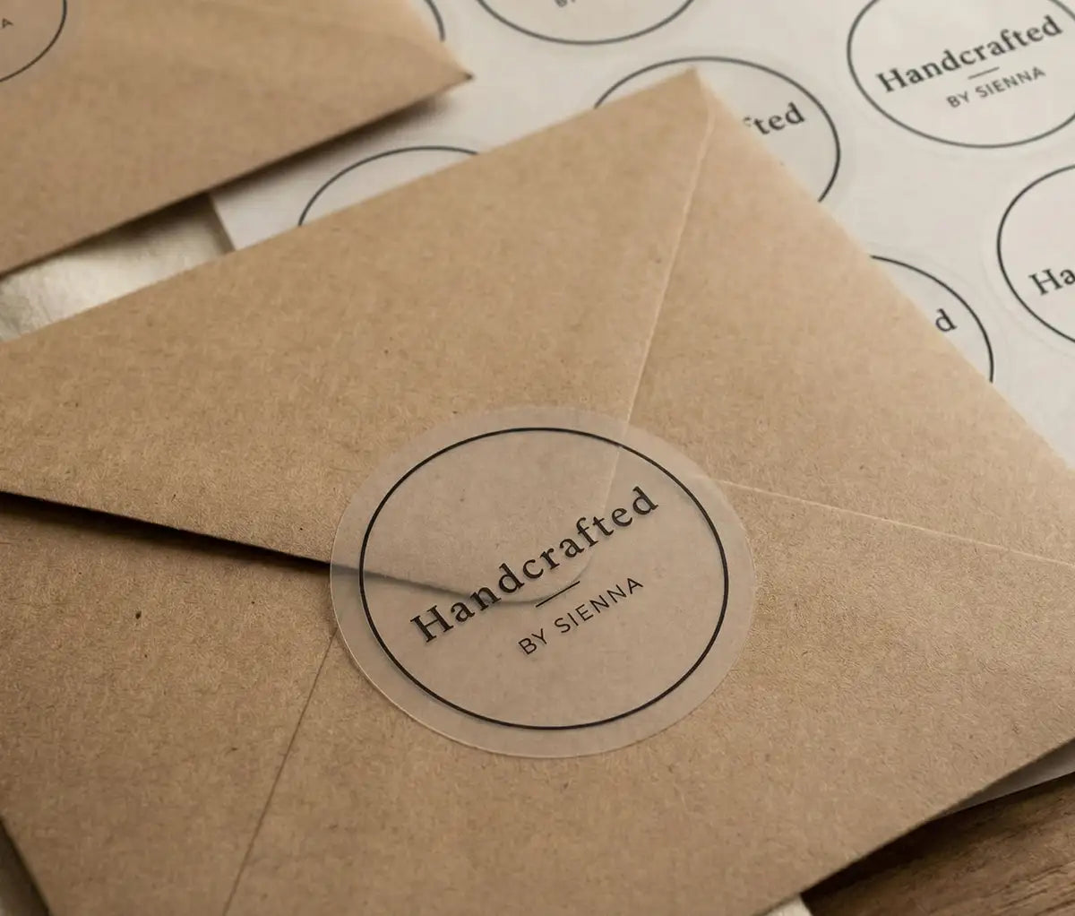 Custom stickers for a small handmade business with text "Handcrafted" and a thin circle frame printed in black ink applied to a kraft colour envelope. There is a sticker sheet behind the envelope.
