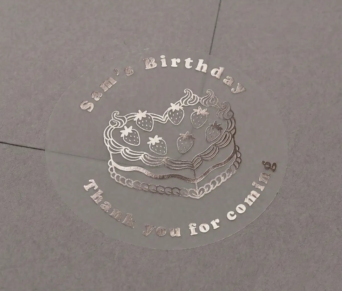 Heart shaped birthday cake, custom name and text "Thank you for coming" printed in silver on transparent sticker that is applied to a grey envelope.