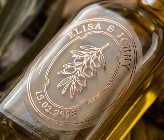 Glass bottle of olive oil with personalized oval frosted sticker featuring olive graphic printed in rose gold foil.