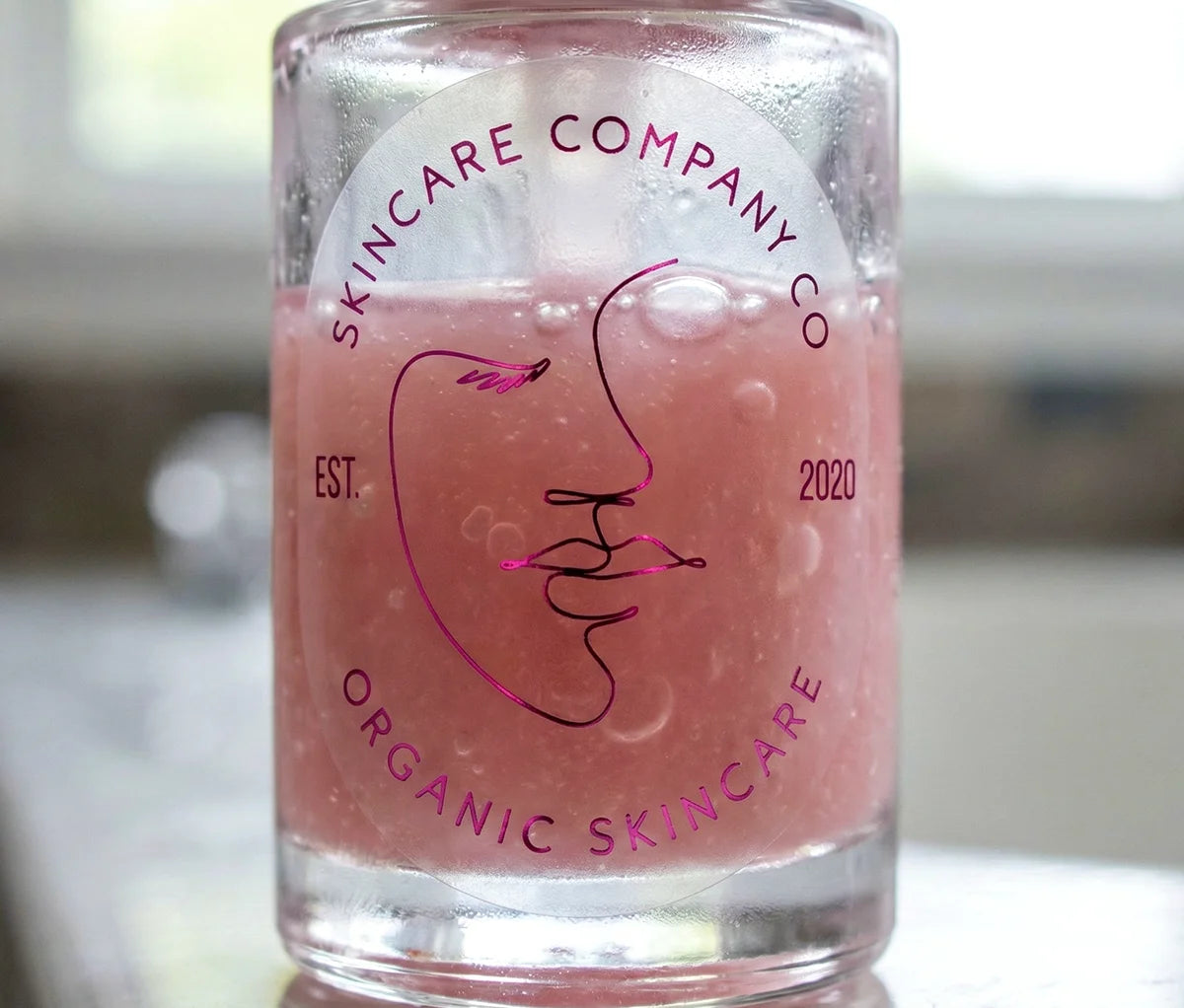 Skincare brand sticker with customizable text on oval frosted sticker printed in Hot Pink Foil and applied to a skincare serum bottle.