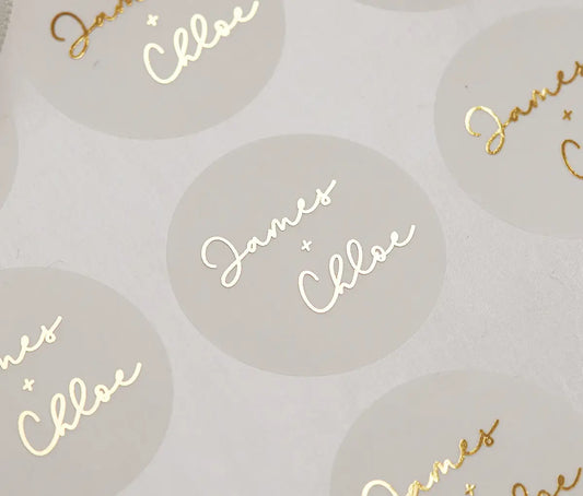 A close-up photograph of frosted sticker sheet of 2 custom names and a "+" sign printed in gold foil.