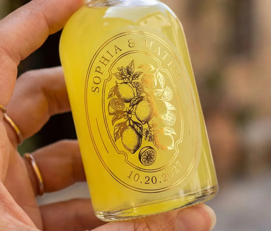 Woman’s hand holding favor sized limoncello bottle with frosted oval label applied to it. Label is printed in gold foil and features couples names, date and lemon graphic.