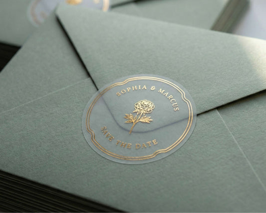 Clear Save the Date sticker with flower graphic and personalized names printed in gold foil sealing sage green envelope.