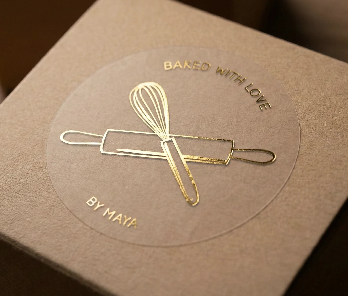 Small kraft box with gold foil printed sticker with an imagage of a whisk and rolling pin, and text 'Baked with Love by Maya'