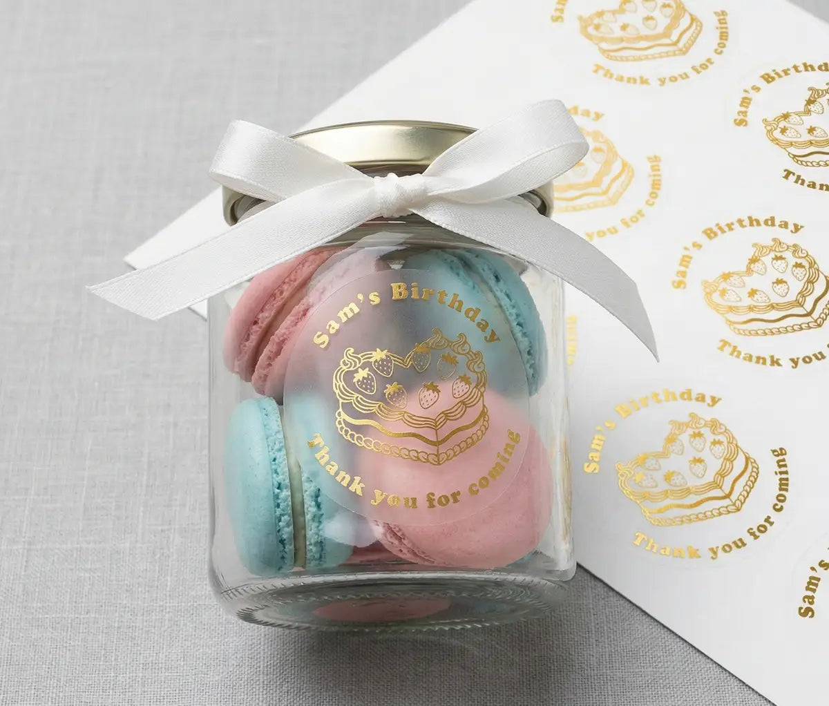 Glass jar with pink and blue macarons and a white ribbon with 'Sam's Birthday' and heart-shaped cake graphic printed in gold foil on frosted sticker applied to the jar. Behind the jar there is a sticker sheet.
