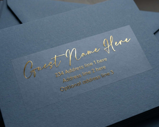 Custom Guest Address Label printed in calligraphy font and in gold foil on transparent matte sticker and applied to a slate blue envelope.