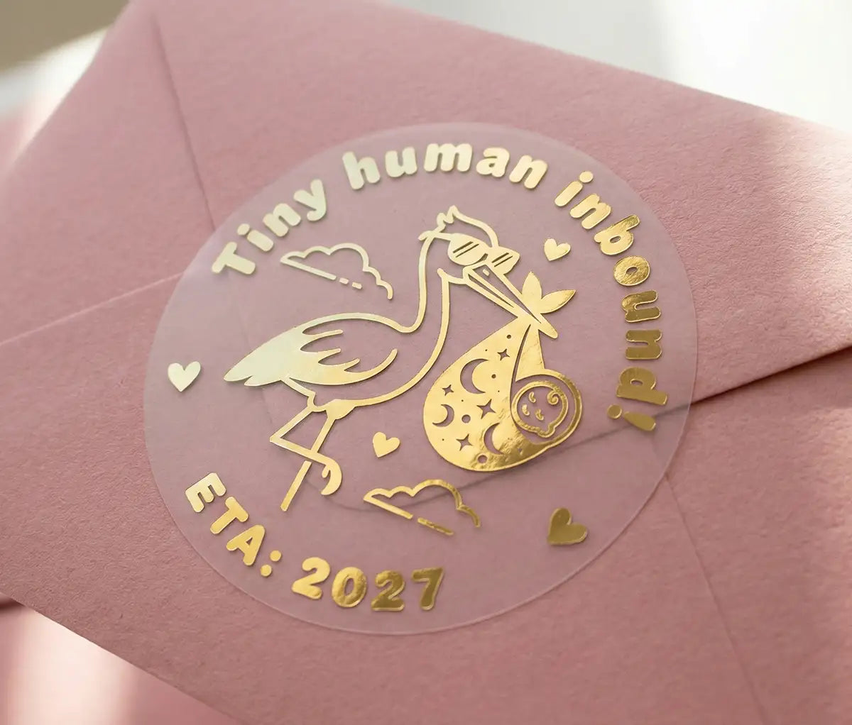Custom baby announcement sticker with real metallic gold foil on a pink envelope. Sticker design shows a stork and the text 'Tiny human inbound! ETA: 2027' printed on a transparent frosted round sticker