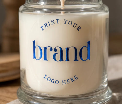 A close-up of a clear round glossy sticker placed on a candle jar, with royal blue foil text reading 'Print Your Logo Here'