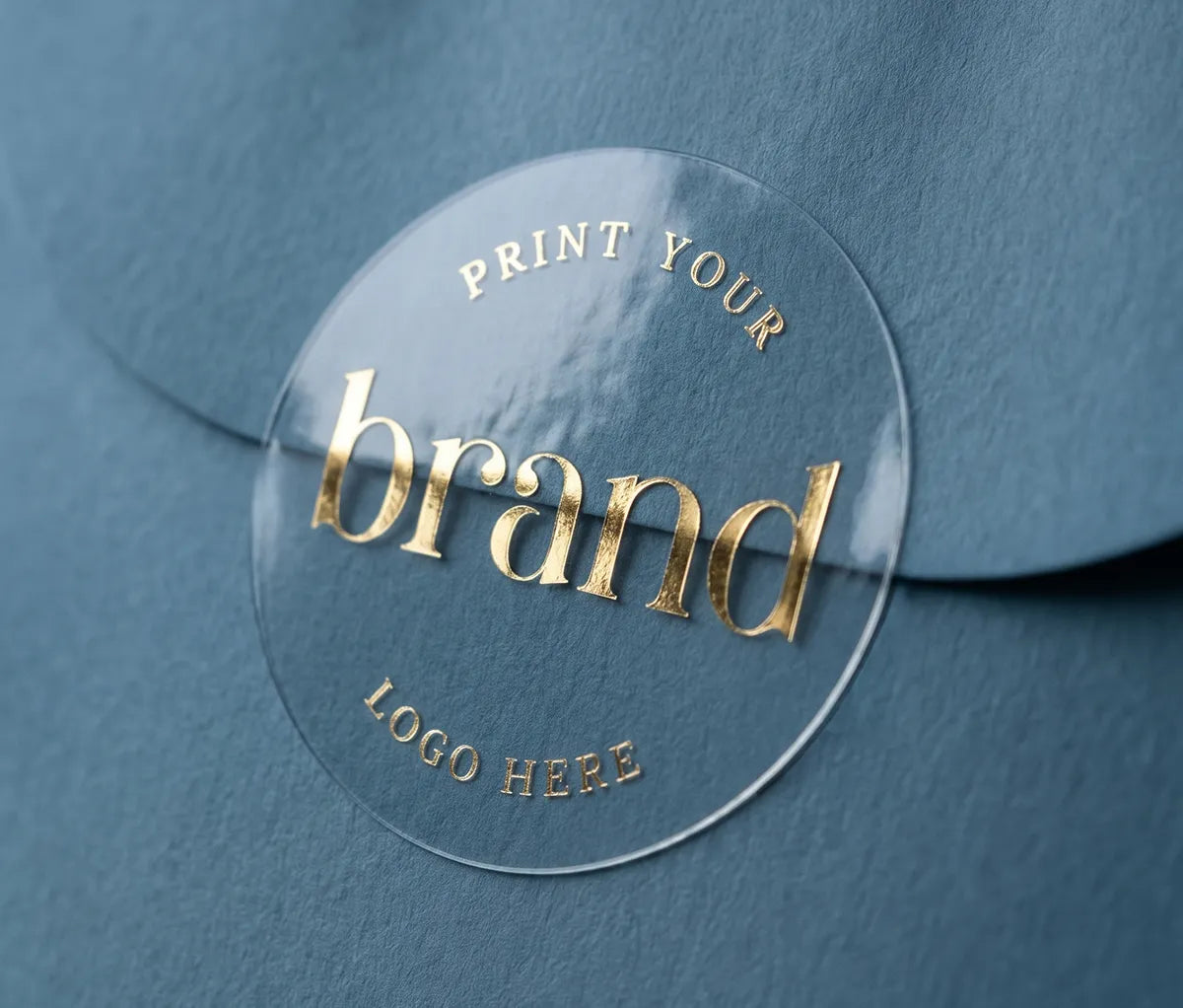A close-up of a clear round glossy sticker placed on a textured blue card, with gold foil text reading 'Print Your Logo Here'
