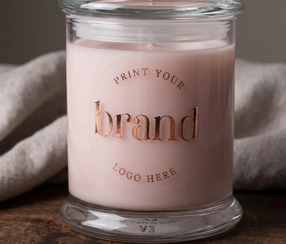 A close-up of a clear round glossy sticker placed on a pink candle jar, with rose gold foil text reading 'Print Your Logo Here'