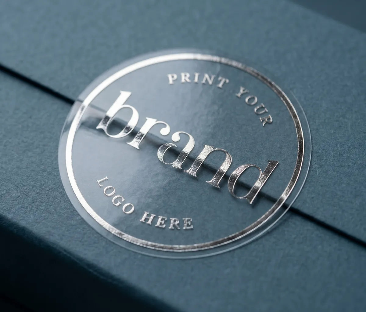 A close-up of a clear round glossy sticker placed on a textured blue card, with silver foil text reading 'Print Your Logo Here'