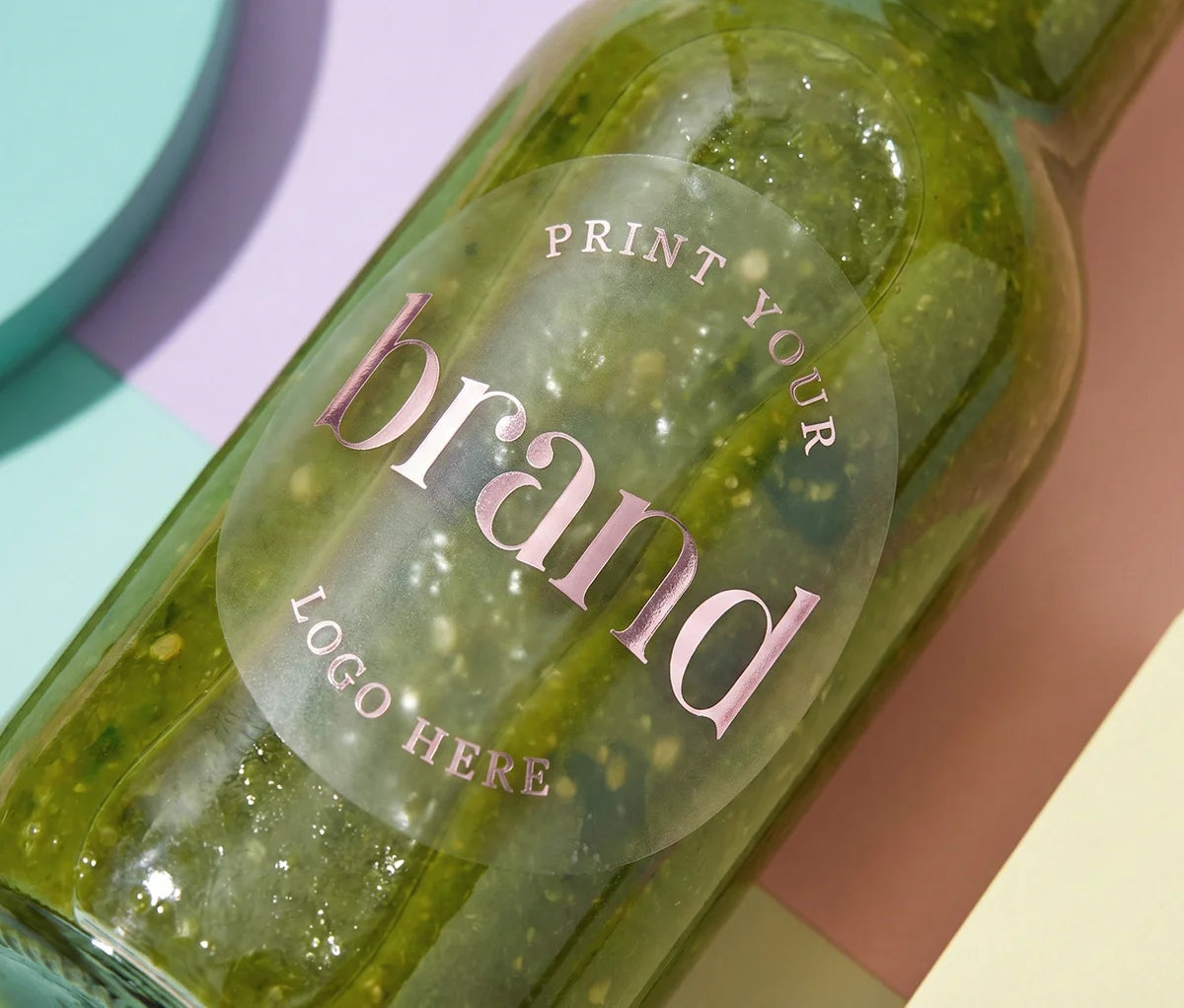Frosted round sticker with custom logo printed in pale pink foil attached to a green sauce glass bottle.
