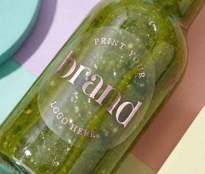 Frosted round sticker with custom logo printed in pale pink foil attached to a green sauce glass bottle.