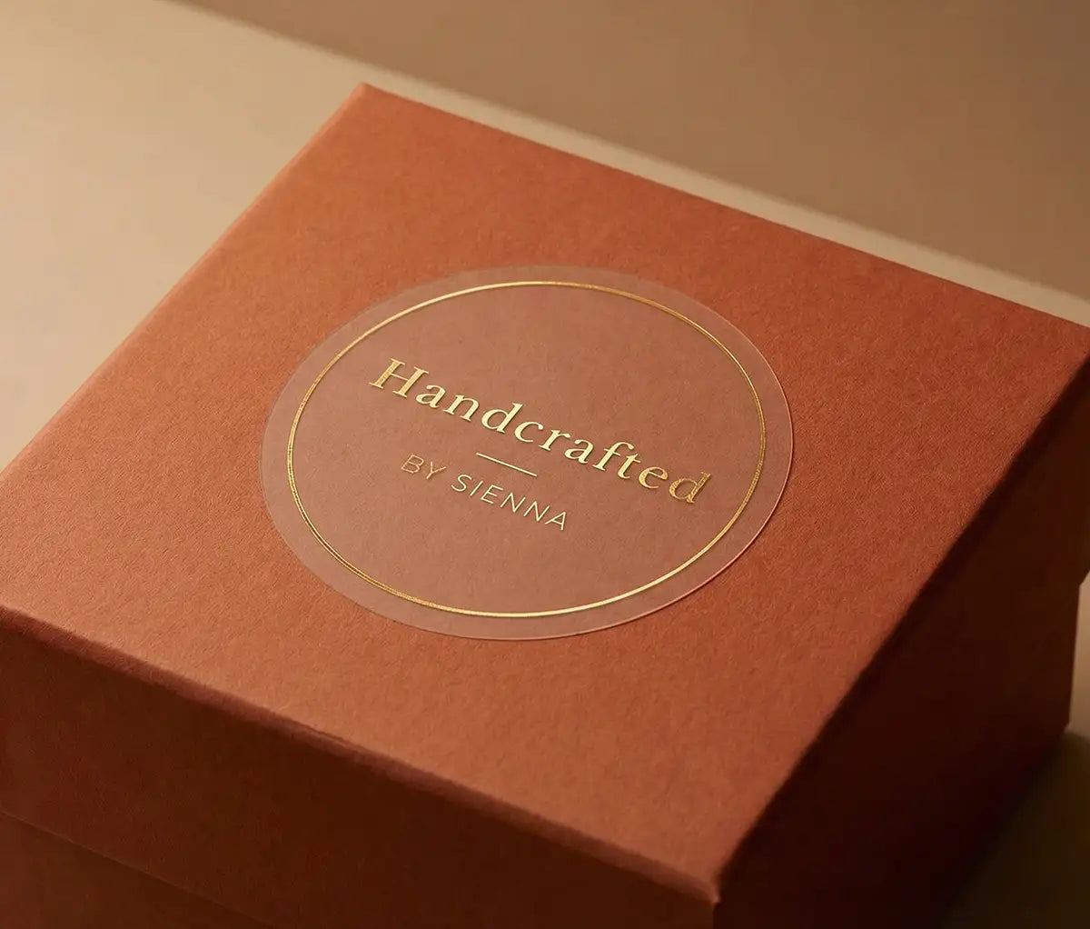 Close-up of transparent sticker with custom text "Handcrafted" and thin circle frame printed in gold foil applied to a terracotta colour gift box. 