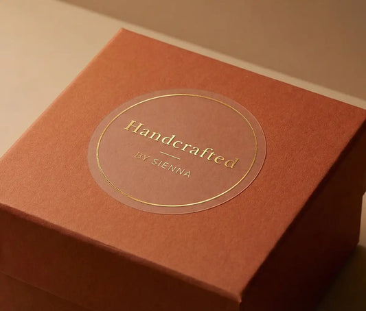 Close-up of transparent sticker with custom text "Handcrafted" and thin circle frame printed in gold foil applied to a terracotta colour gift box. 