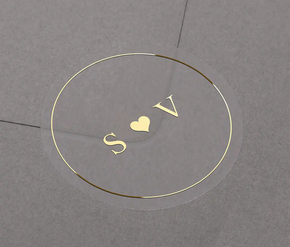 Gold foil wedding sticker with custom initials "S and V" and a small heart centered within it. The label is applied to a grey envelope.