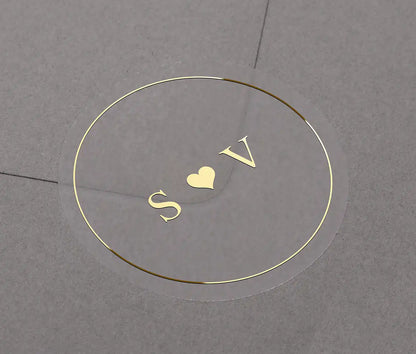 Gold foil wedding sticker with custom initials "S and V" and a small heart centered within it. The label is applied to a grey envelope.