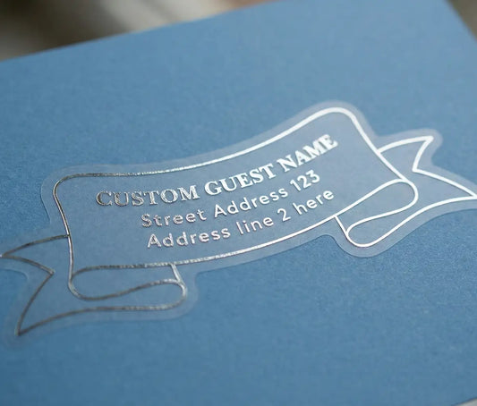 Recipient address label printed in silver foil and custom die cut in unique ribbon shape applied to a blue envelope.