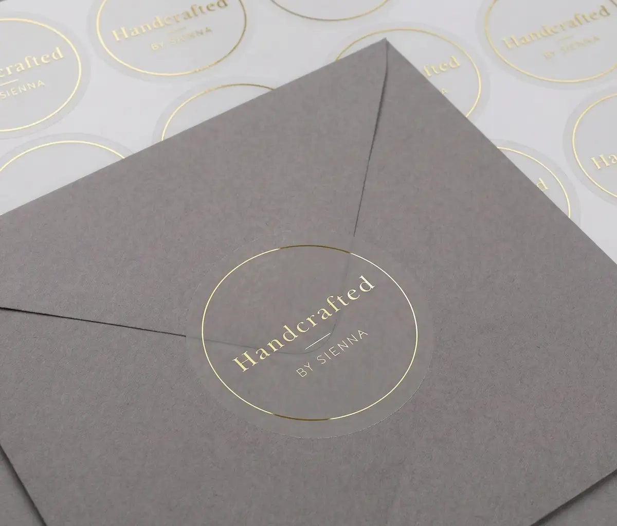 Transparent sticker with custom text "Handcrafted" and thin circle frame printed in goldfoil applied to a grey colour envelope. There is a sticker sheet with the same stickers on white sheet behind the envelope.