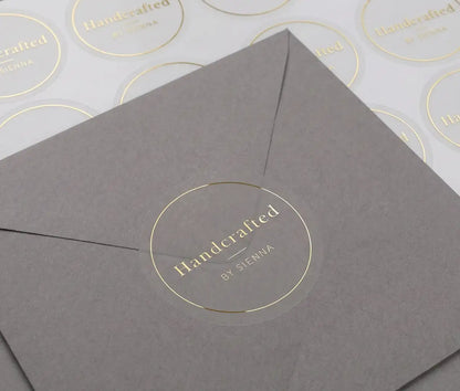 Transparent sticker with custom text "Handcrafted" and thin circle frame printed in goldfoil applied to a grey colour envelope. There is a sticker sheet with the same stickers on white sheet behind the envelope.