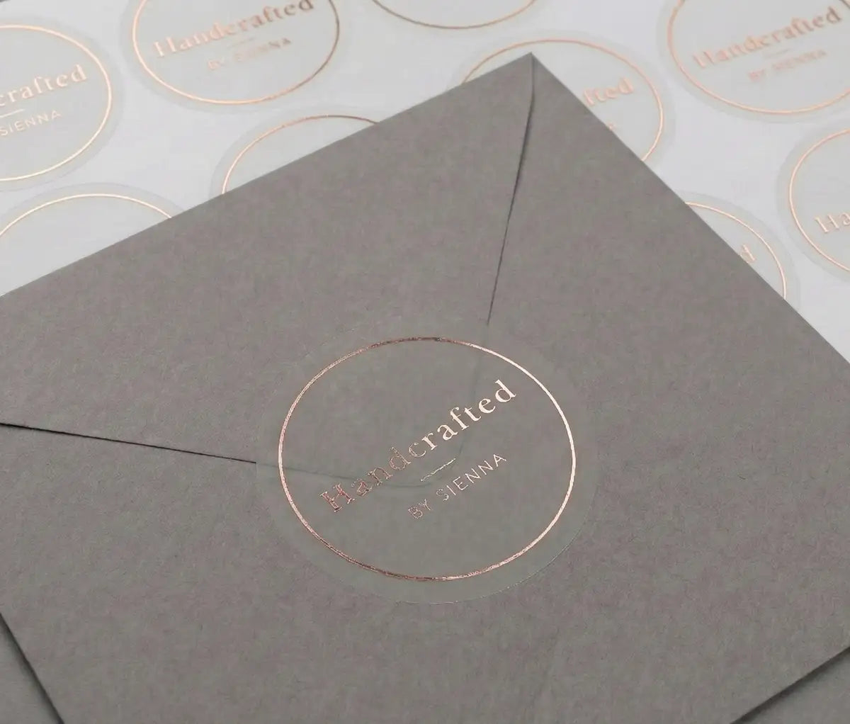Transparent sticker with custom text "Handcrafted" and thin circle frame printed in rose gold foil applied to a grey colour envelope. There is a sticker sheet with the same stickers on white sheet behind the envelope.