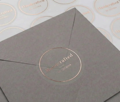 Transparent sticker with custom text "Handcrafted" and thin circle frame printed in rose gold foil applied to a grey colour envelope. There is a sticker sheet with the same stickers on white sheet behind the envelope.