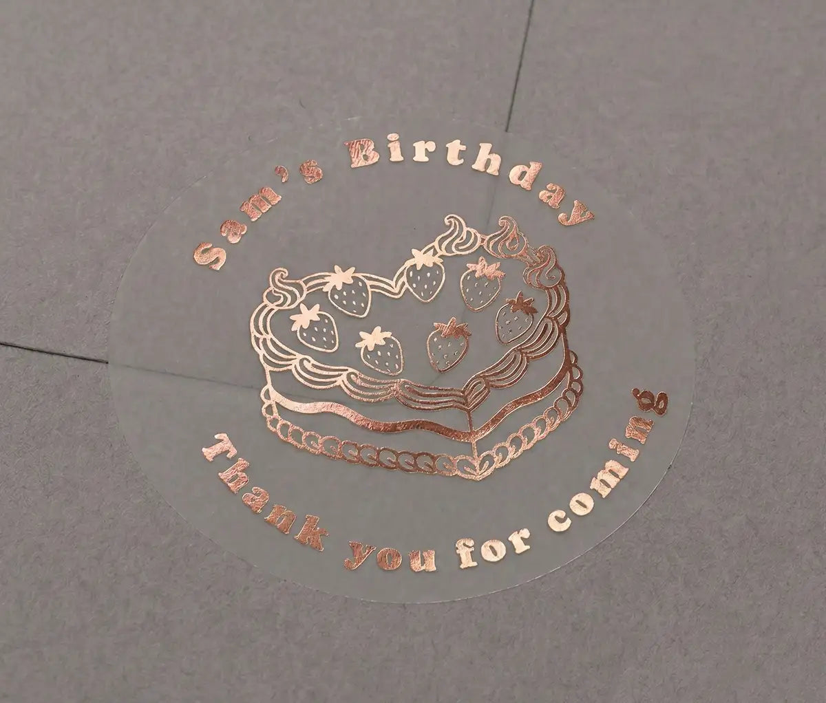Premium rose gold foil-printed birthday stickers with a custom name and text "Thank you for coming" printed on a transparent sticker that is applied to a grey envelope.
