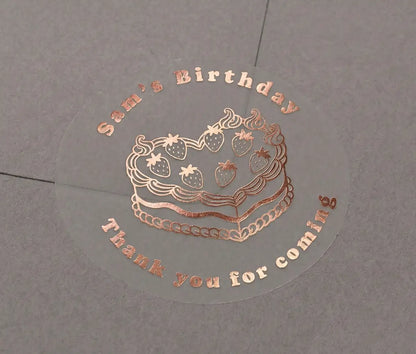 Premium rose gold foil-printed birthday stickers with a custom name and text "Thank you for coming" printed on a transparent sticker that is applied to a grey envelope.