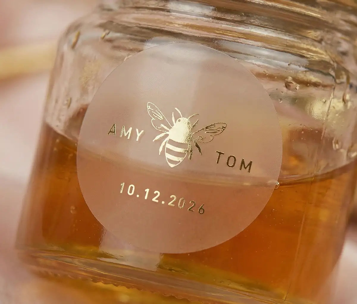 A close-up photograph of a small glass jar filled with golden honey. Affixed to the front is a circular, transparent sticker with gold foil text that reads "AMY TOM" above a graphic of a bee, and date below it. The background is softly blurred with pink and gold tones.