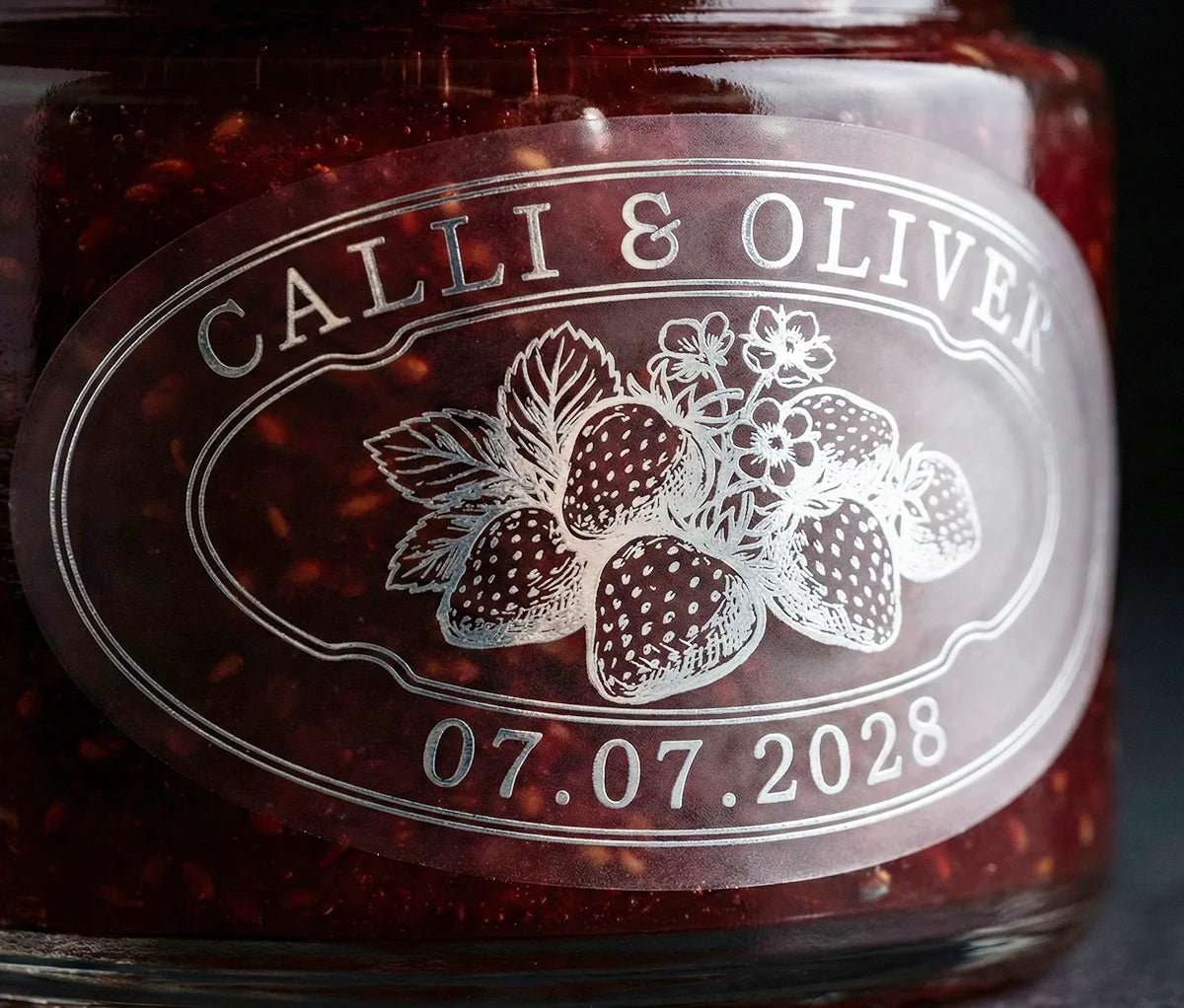 Jar of strawberry jam with personalized oval shape wedding favor sticker printed in silver foil on frosted vinyl.