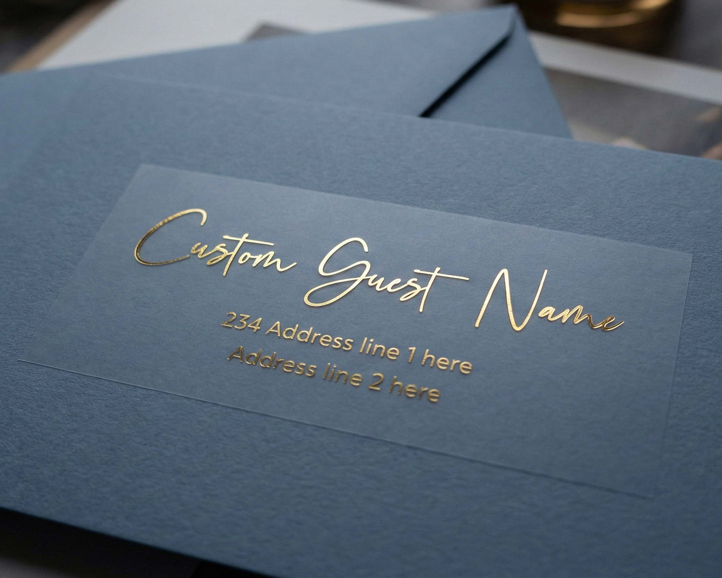 Custom Guest Address Label printed in modern script font and in gold foil on transparent matte sticker and applied to a slate blue envelope.
