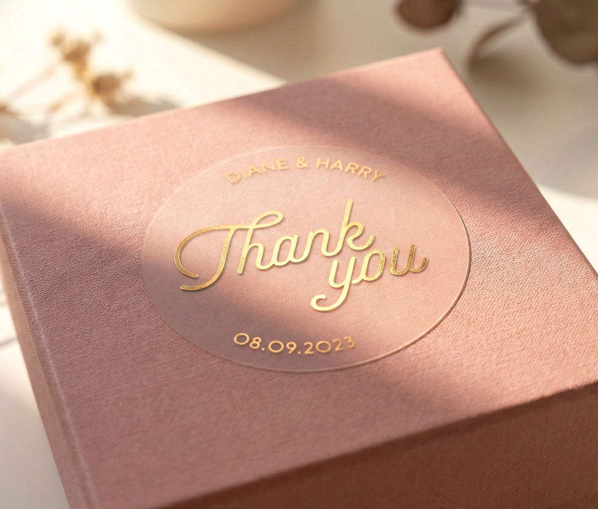 Clear 'Thank you' sticker with personalized names and date printed in gold foil and applied to a pink gift box.