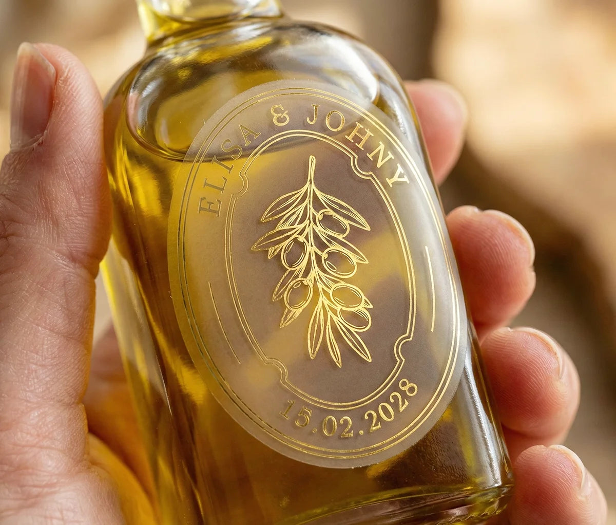 Woman holding glass bottle of olive oil with personalized oval frosted wedding favour sticker printed in gold foil.