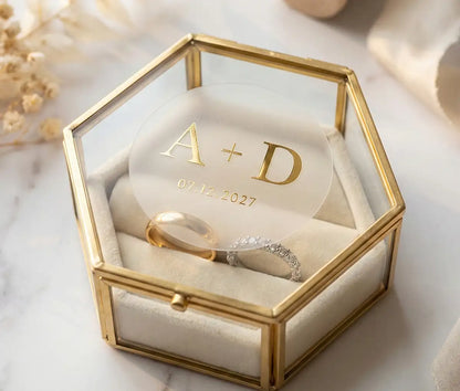 Close-up of a hexagonal glass jewelry box containing wedding rings, featuring a custom round transparent matte sticker on the lid. The design displays initials 'A + D' and a date in real Gold Premium Hot Foil, creating a sleek, smooth finish with a stunning mirror-like shine. The elegant setting includes cream-colored dried florals and ribbon, perfect for high-end wedding stationery inspiration.