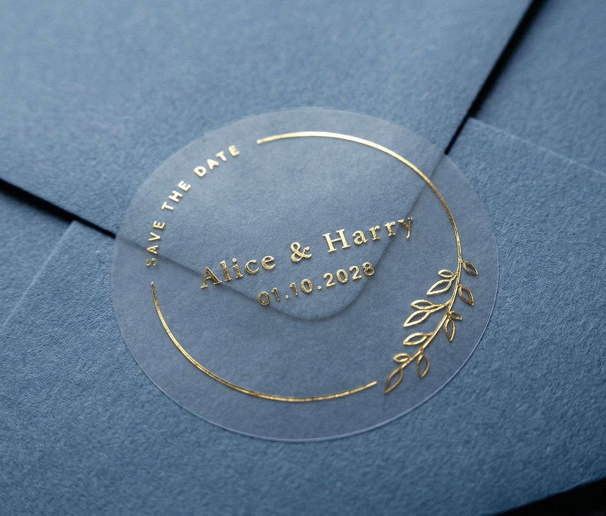 Clear round Save the Date sticker with dainty floral frame and personalized names and date printed in gold foil sealing navy blue envelope.