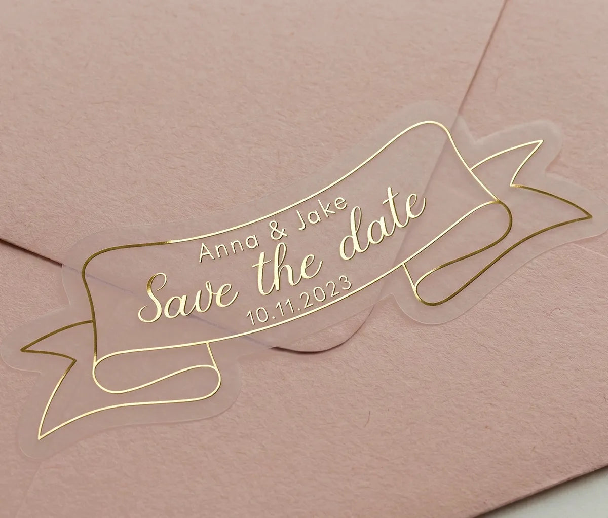 Gold foil printed custom "Save the Date" wedding sticker applied to a pink envelope. Sticker is custom die cut to a ribbon shape.