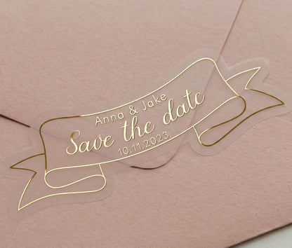 Gold foil printed custom "Save the Date" wedding sticker applied to a pink envelope. Sticker is custom die cut to a ribbon shape.