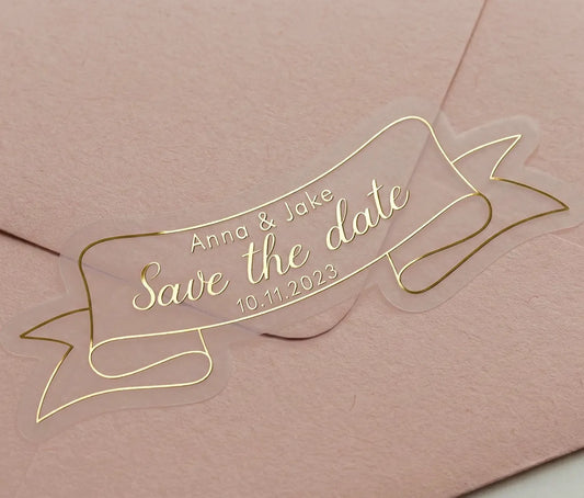 Gold foil printed custom "Save the Date" wedding sticker applied to a pink envelope. Sticker is custom die cut to a ribbon shape.