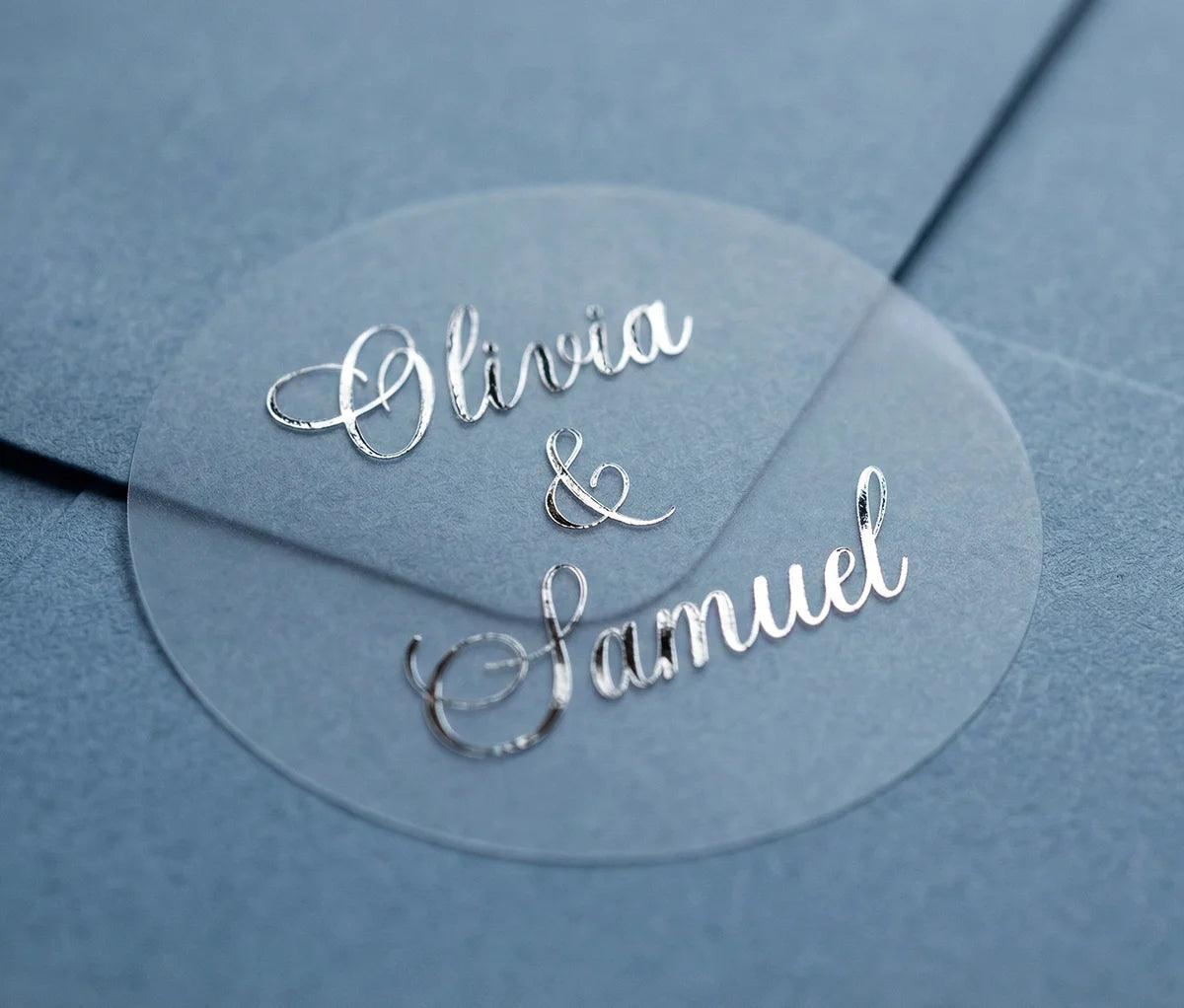 Round transparent wedding sticker with custom names printed in silver foil applied to navy colour envelope.