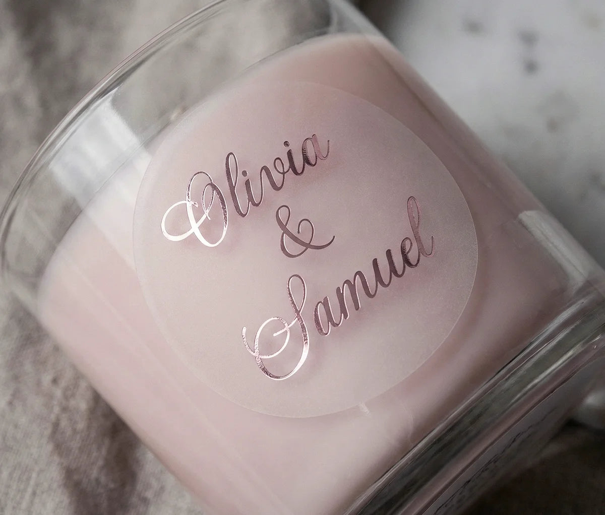 Frosted, round custom wedding sticker with couple's names printed in pale pink foil applied to a glass candle container.