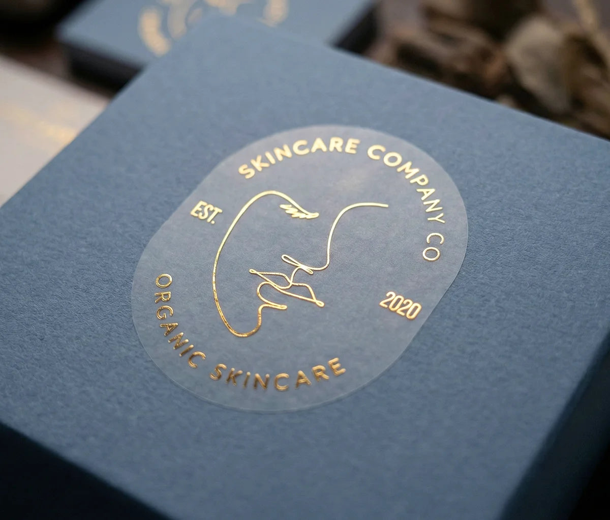 Skincare brand sticker with personalized text on oval frosted sticker printed in Gold Foil and applied to slate blue box.