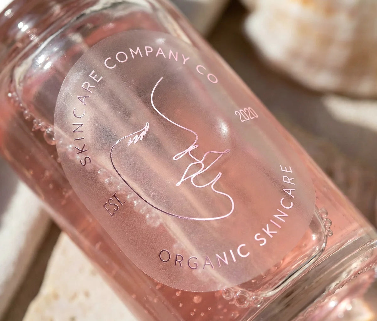 Skincare brand sticker with customizable text on oval frosted sticker printed in Pale Pink Foil and applied to a glass bottle.