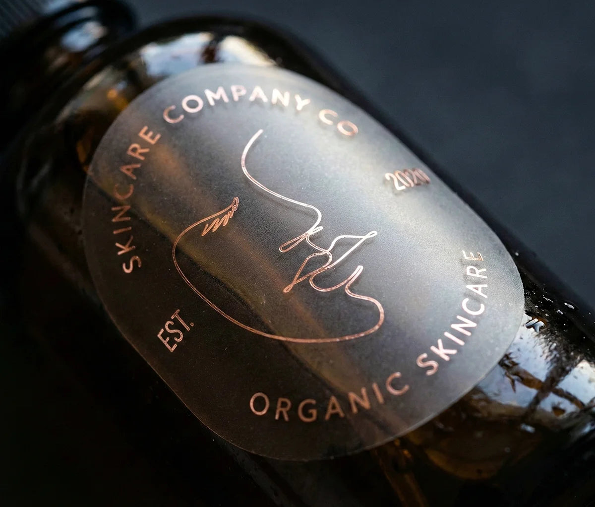 Skincare brand sticker with customizable text on oval frosted sticker printed in Rose Gold Foil and applied to a dark glass bottle.