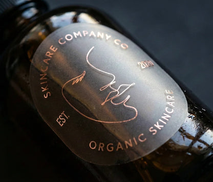 Skincare brand sticker with customizable text on oval frosted sticker printed in Rose Gold Foil and applied to a dark glass bottle.