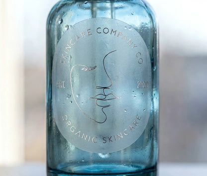 Skincare brand sticker with customizable text on oval frosted sticker printed in Silver Foil and applied to a blue skincare serum bottle.