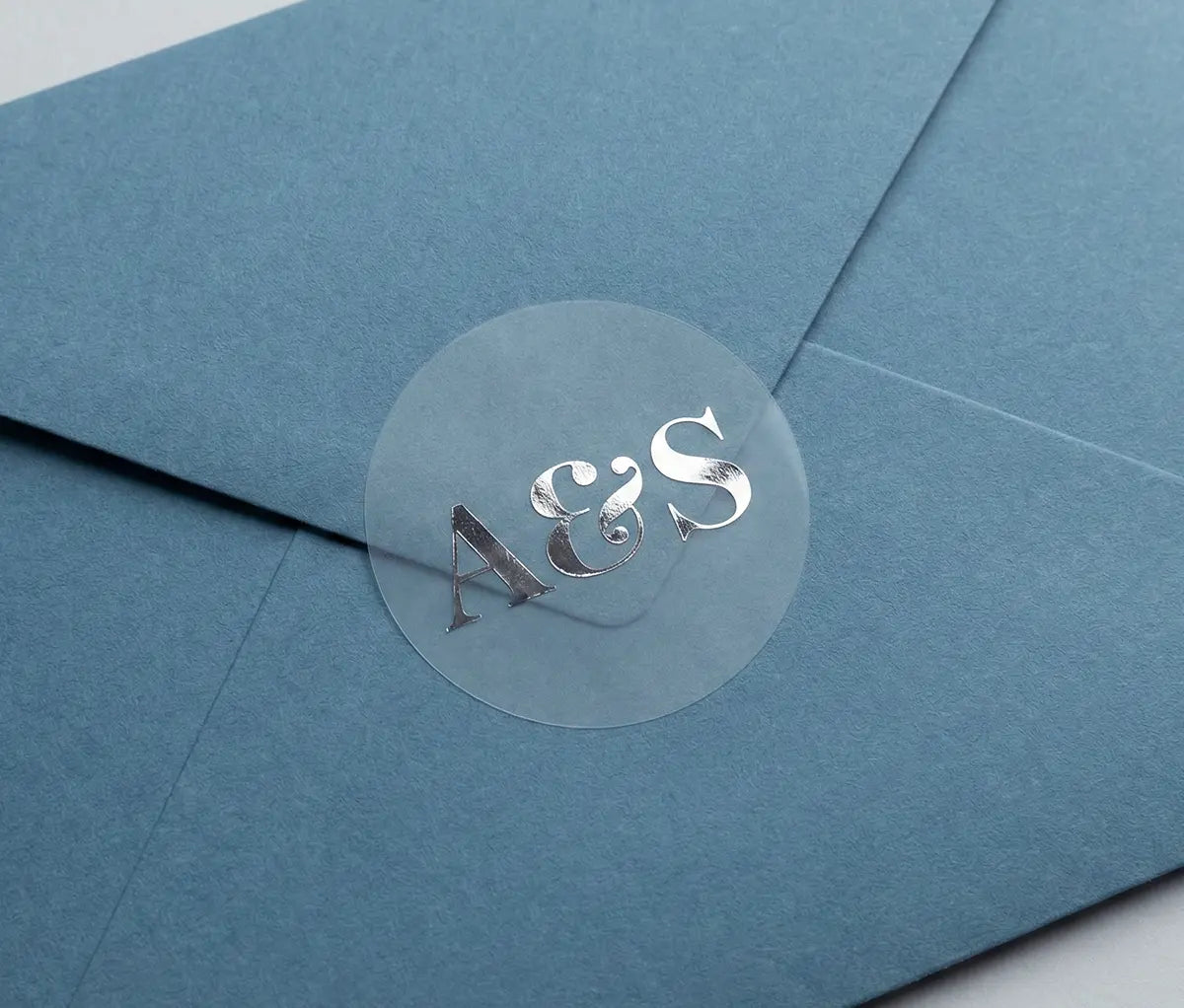 Blue envelope with round Wedding envelope stisker with personalized initials printed in silver foil on transparent matte vinyl.