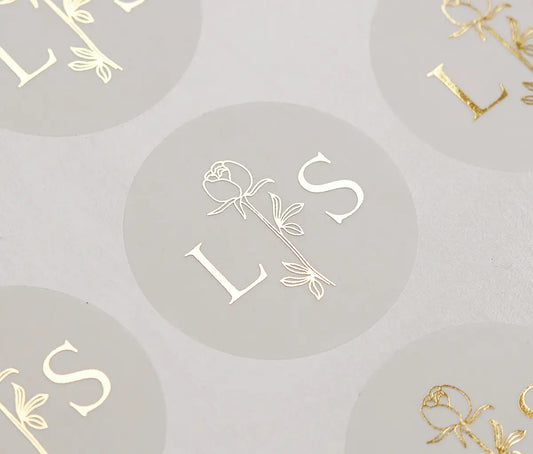 A close-up photograph of gold foil print sticker sheet with custom initials and rose graphic on circle frosted stickers.