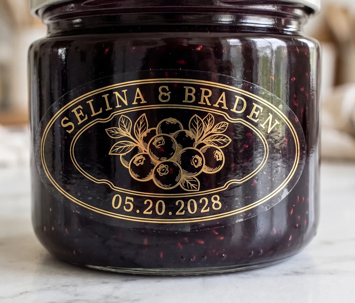 Oval personalized gold foil print clear wedding favour label on a blueberry jam jar.